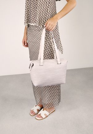 Bolso de mano rosa claro con un acabado texturizado, forma rectangular y dos correas para el hombro, sostenido por una persona que lleva un atuendo estampado y sandalias.