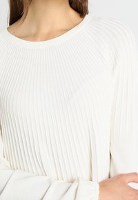 Weißer gerippter Pullover mit rundem Ausschnitt und langen, leicht gepufften Ärmeln mit gerafften Manschetten. Weiche Stricktextur.