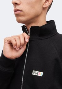 Άνδρας κλείνει μαύρο μπουφάν με ψηλό γιακά· το μπουφάν έχει ένα μικρό λογότυπο "JUUE" με μπλε, ροζ και κίτρινες ρίγες στο στήθος.