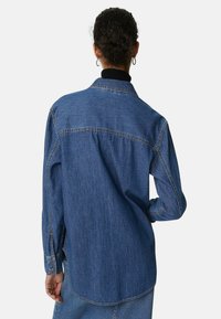 Denimskjorta i mörkblått med krage, lång ärm och knappar vid ärmsluten. Har en avslappnad passform och synliga sömnadsdetaljer.