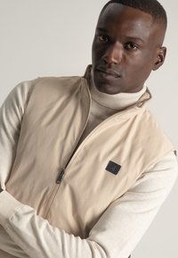 Chaleco sin mangas beige con un alto cuello, cremallera delantera y un pequeño parche negro con logo en el pecho. Llevarlo sobre un cuello de tortuga de color claro.
