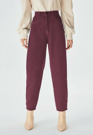 Femme portant un pantalon en velours côtelé bordeaux taille haute, un pull beige en tricot et des bottines beiges à bout pointu sur un fond clair uni.