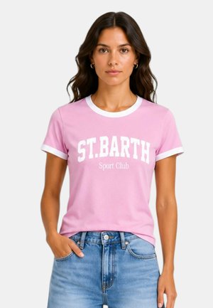 Donna con capelli scuri e mossi che indossa una T-shirt rosa con bordo bianco con la scritta "ST.BARTH Sport Club", abbinata a jeans blu, posa con una mano in tasca.