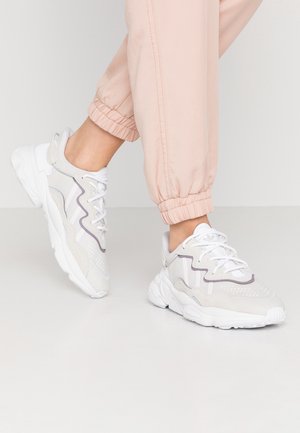 Osoba nosząca białe i szare sneakersy z białymi sznurowadłami i jasnoróżowe spodnie dresowe stojąca na białej podłodze.