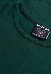 Grøn bomulds t-shirt med rund halsudskæring og en syet kant. Indeholder et "Superdry Athletic Essentials" label på siden.