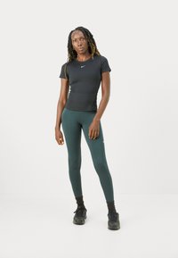 Idrettskvinne med langt flettehår iført svart tettsittende Nike T-skjorte, turkise leggings, svarte strømper og svarte joggesko som står mot en hvit bakgrunn.
