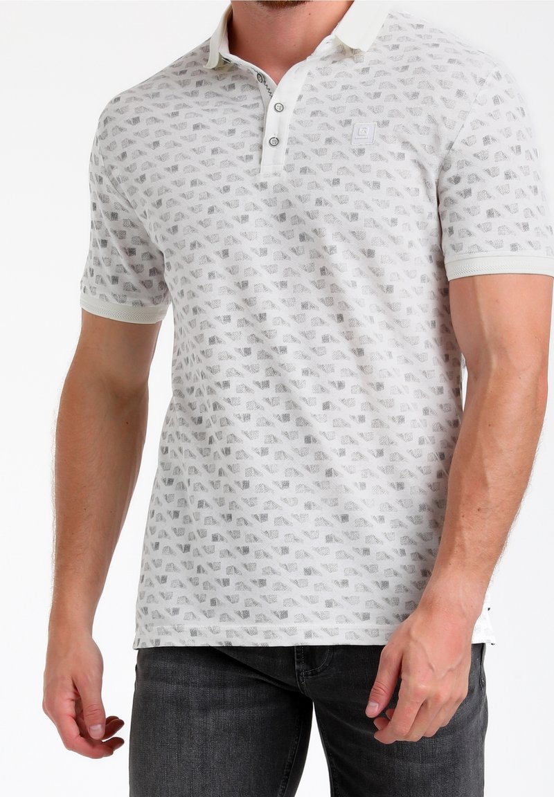 Homme portant un polo blanc à motifs à manches courtes et un jean foncé, debout devant un fond blanc.