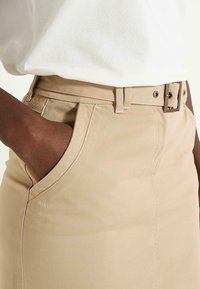 Jupe en coton beige avec une taille ajustée, des poches latérales et une ceinture avec une boucle carrée, présentée avec une main dans une poche.