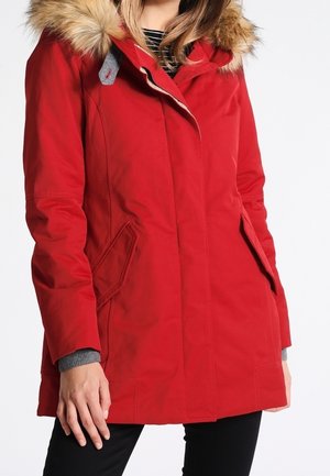 Woman wearing a red winter coat with fur-trimmed hood, side pockets, and gray sweater cuffs, standing with one hand relaxed.