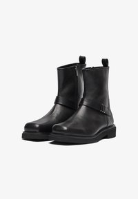 Botas de tobillo de cuero negro con una textura suave, que presentan una cremallera lateral, una correa con hebilla y una suela de goma resistente.