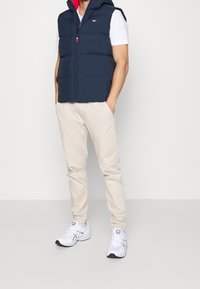 Granatowa kamizelka puchowa z kapturem, posiada czerwoną podszewkę i zamek błyskawiczny z przodu; zestawiona z jasnobeżowymi spodniami i białymi butami sportowymi.