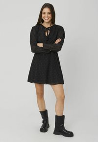 Robe noire en A avec manches longues, décolleté à nouer et tissu texturé. Associée à des bottines noires épaisses.