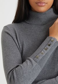 Grauer Rollkragenpullover aus weichem Stoff, mit enganliegendem Design und dekorativen silbernen Knopfakzenten an den Manschetten.