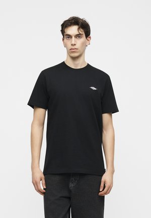 Jeune homme aux cheveux courts et foncés portant un t-shirt noir avec un petit logo blanc sur la poitrine et un jean foncé, face à un fond blanc.