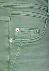 Tela de denim verde con detalles de costura visibles; presenta un pequeño bolsillo frontal asegurado por herrajes plateados y costuras de acento en verde.