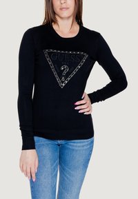 Maglione nero a maniche lunghe con un design a logo triangolare argentato. Polsini e orlo a coste, realizzato in tessuto morbido, indossato con jeans azzurri chiari.