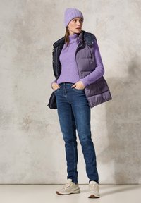 Pull en tricot violet, gilet bleu marine, jeans bleu foncé et baskets claires portés par un mannequin posé sur un fond texturé.