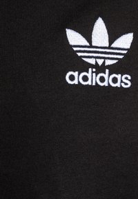 Tessuto di cotone nero con un logo Adidas ricamato bianco, arricchito da tre dettagli a forma di foglia e tre strisce orizzontali sotto il logo.