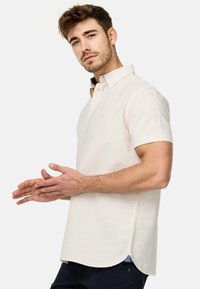 Camisa polo de manga corta beige con cuello, que presenta un forro interior azul oscuro y aberturas laterales. Textura suave con un sutil detalle de logo.