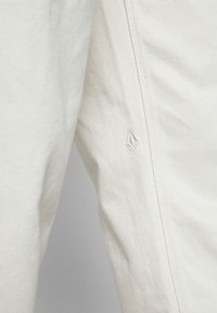 Gros plan sur un tissu blanc avec une couture visible et un petit logo brodé en forme de feuille géométrique.