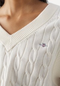 Gros plan d'une personne portant un pull crème à col en V avec des torsades, orné d'un petit blason brodé sur la poitrine.