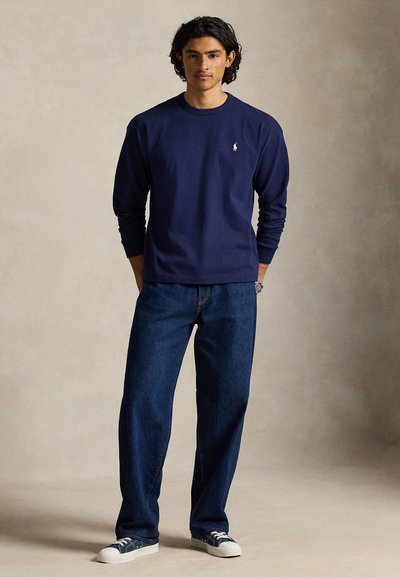 Νέος άνδρας στέκεται με τα χέρια στις τσέπες, φοράει μπλε σκούρο μακρυμάνικο πουκάμισο, άνετο μπλε τζιν και μπλε αθλητικά παπούτσια μπροστά σε απλό φόντο.