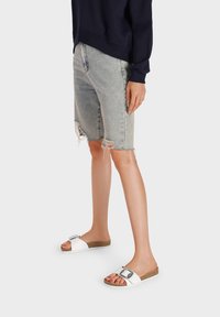 Infradito bianchi con una fibbia metallica, plantare color tan e suola textured. Abbinati a jeans corti in denim leggermente rovinati di colore blu chiaro.