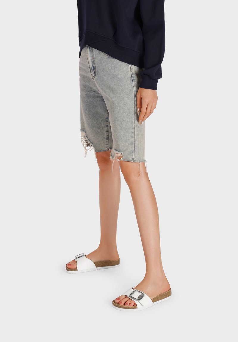 Infradito bianchi con una fibbia metallica, plantare color tan e suola textured. Abbinati a jeans corti in denim leggermente rovinati di colore blu chiaro.