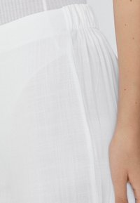 Weißer, leichter Stoff mit einem gerafften Bund, der eine subtile Textur und glatte Oberfläche aufweist. Nahaufnahme des seitlichen Details des Kleidungsstücks.