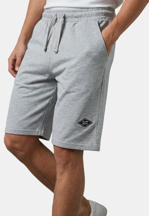Homme portant un short gris clair à cordon avec des poches latérales et un petit logo noir en forme de diamant sur la jambe gauche.