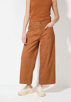 Braune weite Hosen aus Baumwolle, mit zwei vorderen Taschen, einer hohen Taille mit zwei Knöpfen und einer glatten Textur. Tragen mit weißen Sneakers.