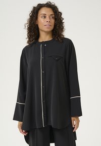 Melnā pogā pārgā krekliņā, izgatavots no gludas auduma. Tam ir vērsta apkakle, krūšu kabata, balta apmale gar priekšpusi un piedurknēm. Garas piedurknes.