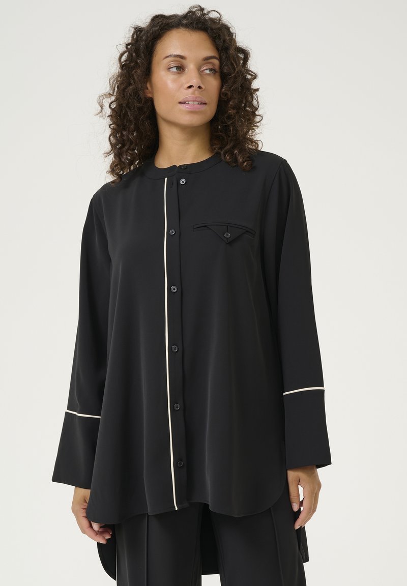 Melnā pogā pārgā krekliņā, izgatavots no gludas auduma. Tam ir vērsta apkakle, krūšu kabata, balta apmale gar priekšpusi un piedurknēm. Garas piedurknes.