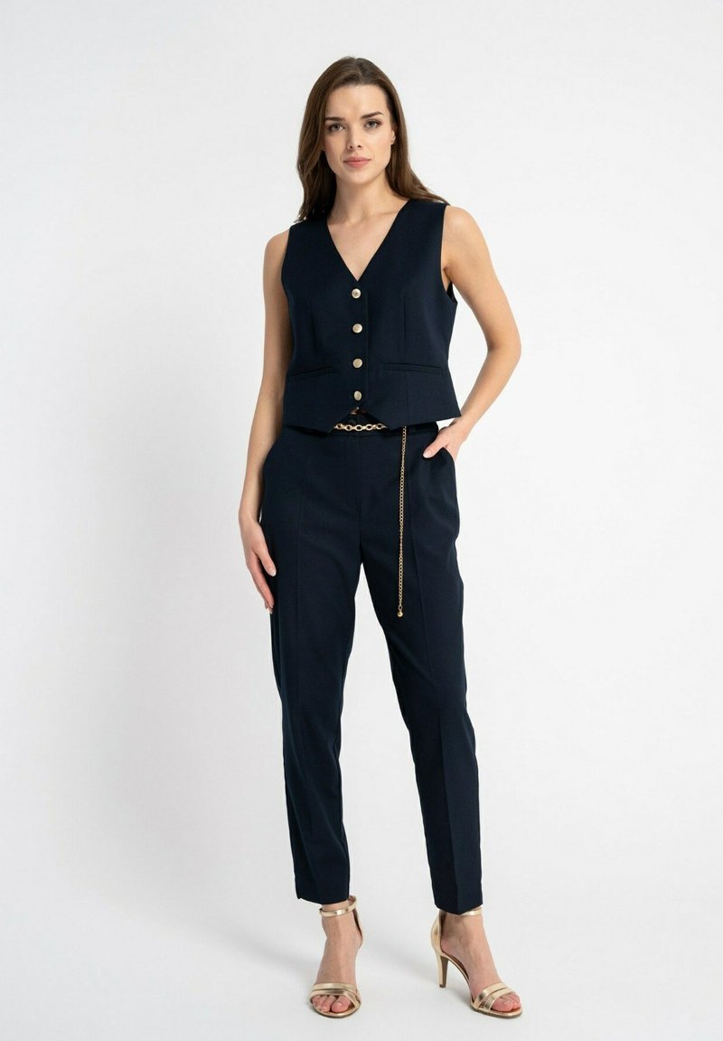 Granatowa kamizelka z dekoltem w serek, złotymi guzikami i dwoma kieszeniami, zestawiona z dopasowanymi spodniami i metalicznymi łańcuchowymi akcentami.