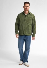Olive green jacket in a sturdy fabric with a collar, button front, and two front pockets. Worn with blue jeans and white sneakers.