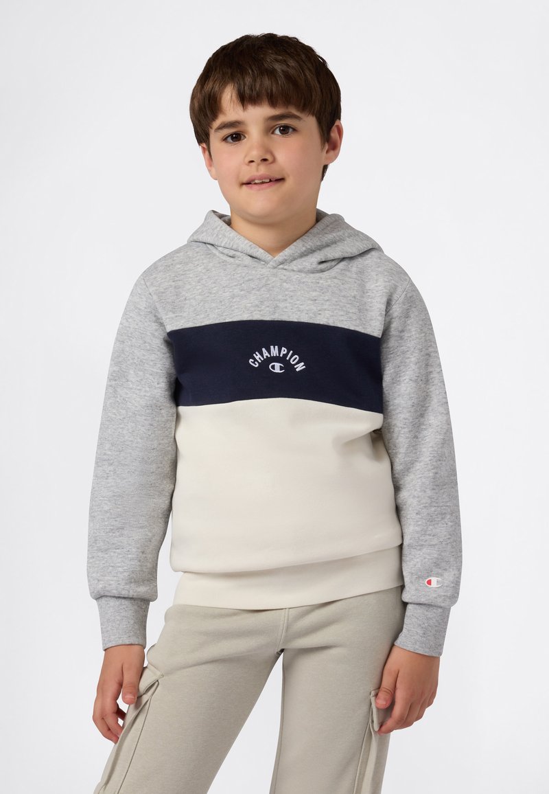 Chico vestido con una sudadera Champion gris, azul marino y crema, con pantalones cargo beige, de pie con las manos relajadas a los lados frente a un fondo liso.