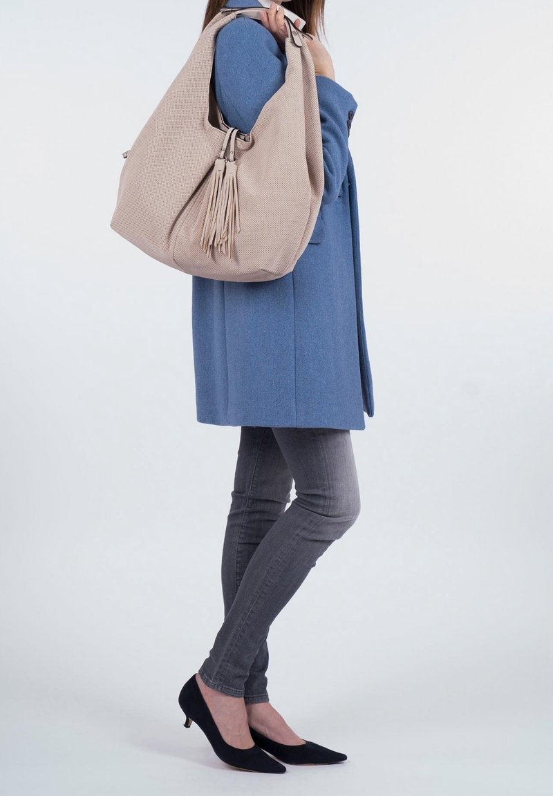 Personne portant un manteau bleu, un jean skinny gris, des talons noirs, tenant un grand sac à bandoulière beige avec un détail pompon.