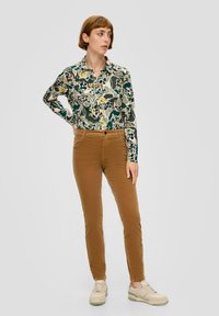 Bloemenoverhemd met knopen in groen, geel en witte patronen; gecombineerd met bruine corduroy broek en beige sneakers.