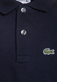 Polo shirt blu navy realizzato in tessuto testurizzato, con colletto, due bottoni in metallo argentato e un logo di coccodrillo verde ricamato sul lato sinistro del petto.