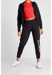Sort zip-up hættetrøje, rød t-shirt med "CALVIN KLEIN" logo, sorte joggerbukser med side-logo. Sorte atletiske sko fuldender outfittet.