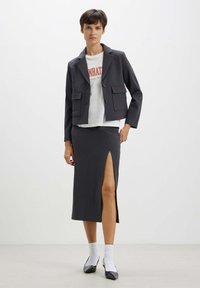 Blazer gris foncé avec deux poches avant, associé à une jupe longue avec une fente latérale. Un T-shirt graphique blanc et des chaussures noires complètent le look.