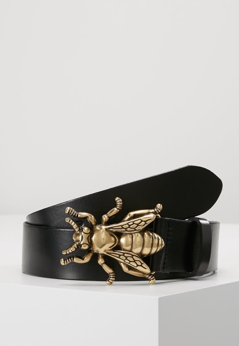 Ceinture en cuir noire avec une grande boucle dorée en forme d'abeille reposant sur une surface blanche contre un fond clair uni.