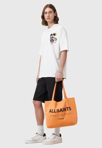 Vit T-shirt med palmtryck och svarta shorts. Klarröd tote-väska med "ALLSAINTS" i svart text. Vita sneakers av bomullstextur.