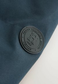 Țesătură de culoare bleumarin cu o suprafață texturată prezintă o emblema rotundă de culoare neagră, împ embossed cu textul "Finest Heritage Brand 1992" și cu un "H" stilizat.