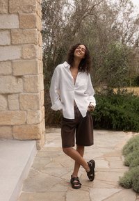 White linen button-up shirt, brown denim shorts, and black sandals. Natural stone wall and greenery in the background.