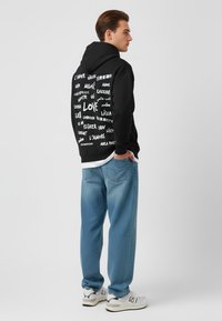 Felpa nera con design di testo bianco raffigurante la parola "AMORE" e varie traduzioni. Abbinata a jeans blu chiaro e sneakers bianche.