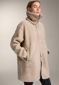 Beige fleece jas met hoge kraag, ritssluiting aan de voorkant en zijzakken. Losse pasvorm met gestructureerd oppervlak en contrasterende bies bij de manchetten.