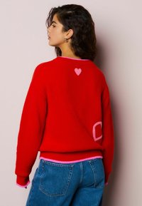 Rode gebreide trui met roze accenten bij de halslijn en manchetten. Heeft een roze hart en de letter 'C' op de achterkant als detail.