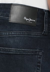 Tył ciemnoniebieskich dżinsów z denimu z czarną skórzaną naszywką marki z napisem "Pepe Jeans London" nad prawą tylną kieszenią.
