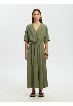 Kvinde stående iført en olivengrøn knap-midi kjole med bånd i taljen, sorte sandaler og to armbånd på hendes højre håndled.